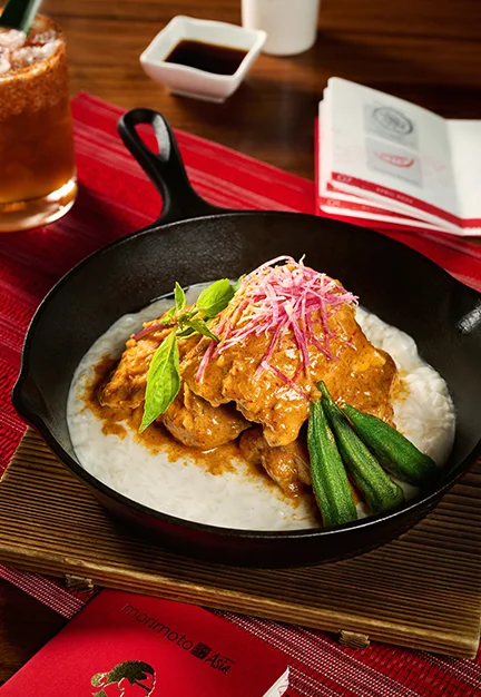 Skillet with chicken curry over white rice topped with shredded pink and white garnish and basil, okra on side, on red placemat beside drink, soy sauce dish, menu; card text reads “Morimoto Asia”.