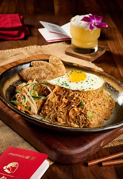 Plate of fried rice topped with a sunny-side egg sits with cucumber salad and crackers on a wooden table beside chopsticks, a flower-garnished cocktail, and a card reading “morimoto”.