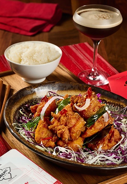 Fried chicken pieces rest on shredded cabbage in a bowl, served with white rice and chopsticks on a wooden table beside a dark cocktail and red napkins in a dining setting