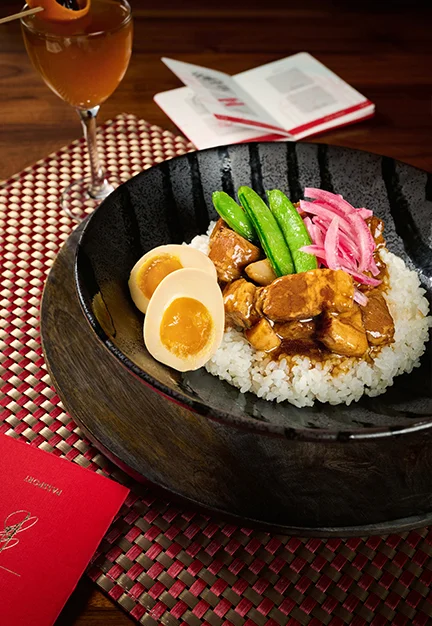 Bowl of white rice topped with braised pork cubes, halved soft-boiled egg, green snap peas, and pickled red onions; cocktail glass, open menu, and card reading “Morimoto Asia” on table.