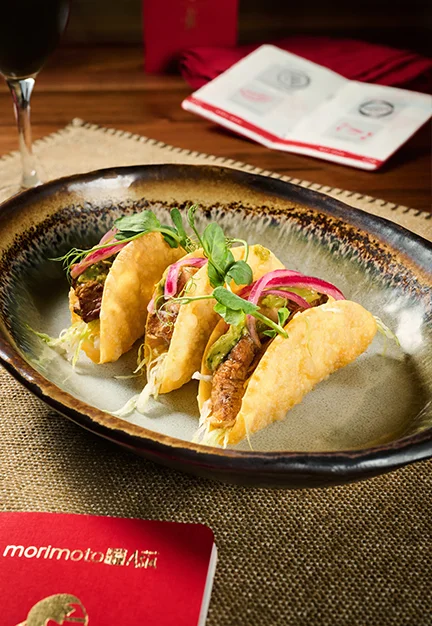 Three tacos filled with sliced beef, shredded cabbage, pickled red onions, and microgreens arranged in a ceramic bowl on a burlap placemat; wine glass, open menu, and card reading “Morimoto Asia” nearby.