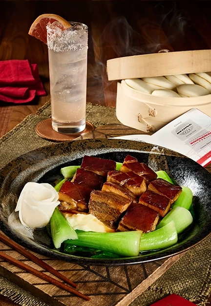 Glazed pork belly pieces sit on green vegetables and white sauce in a bowl, with chopsticks, a tall salted cocktail, and a steaming basket of buns on a wooden table.